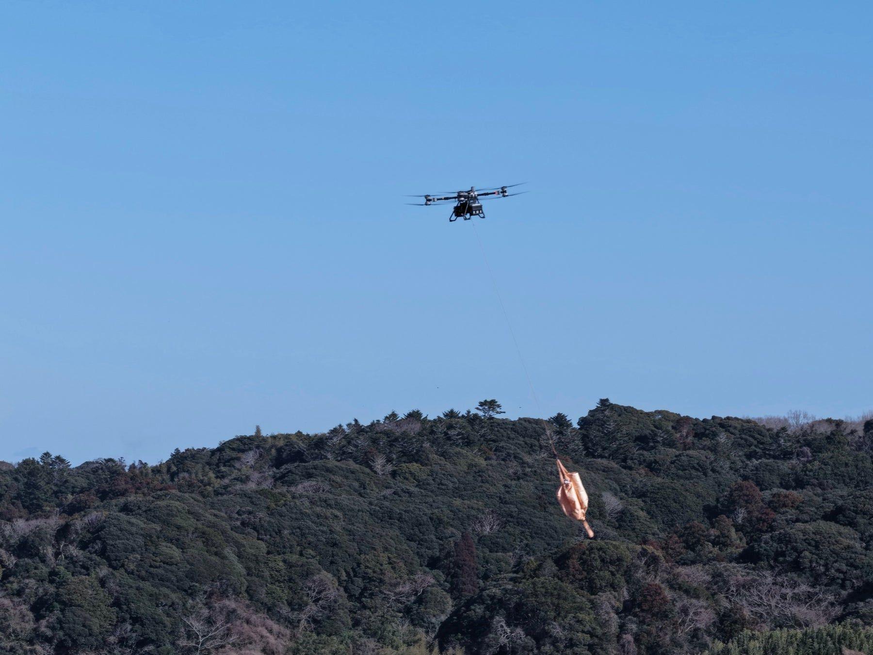 山林の上空を、荷物を吊り下げて飛行する大型物流ドローン